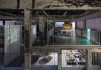 Blick ins Schaudepot des Ruhr Museums auf der Kokerei Zollverein: Zwischen Betonstützen und Metallgeländern sind auf mehreren Ebenen Sammlungsobjekte sowie Videoprojektionen und beleuchtete Elemente einer künstlerischen Installation zu sehen.