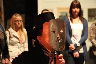 Ein Mensch mit einer Theatermaske wird von anderen Besucher*innen beobachtet.