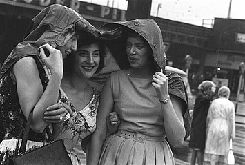 Das Foto von Pressefotografin Marga Kingler zeigt drei fröhliche Frauen, die gemeinsam Schutz unter einer Jacke vor dem Sommerregen im August 1961 in Essen suchen.