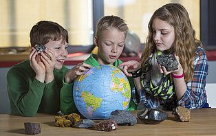 Drei Kinder betrachten einen Globus und halten Steine in der Hand.