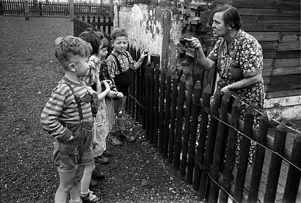 Five children, some in lederhosen, stand at a fence and get a lecture from a neighbor behind it.