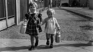 Schwarz-Weiß-Bild von zwei kleinen Kinder, die mit ihren Milchkannen auf dem Bürgersteig laufen.