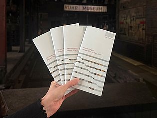 Eine Hand hält vier aufgefächerte Exemplare des neuen Veranstaltungsprogramms des Ruhr Museums für Oktober 2026 bis März 2027. Im Hintergrund ist unscharf der Eingangsbereich des Ruhr Museums zu sehen.