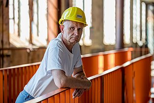 Frank Arnold trägt ein weißes T-Shirt und einen gelben Bauhelm und lehnt auf einem Geländer in seiner alten und neuen Arbeitstelle, der ehemaligen Energiefabrik Knappenrode