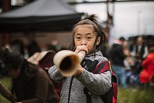 Ein Mädchen mit grauer Jacke und rotem Rucksack bläst konzentriert in ein langes Holzblasinstrument. Im Hintergrund eines belebten Festes sitzen und stehen unscharf weitere Menschen, wodurch die Szene lebendig und gemeinschaftlich wirkt.