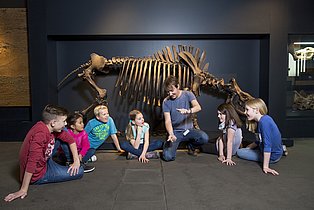 Kinder sitzen gemeinsam mit einem Gästeführer in der Dauerausstellung vor einem großen Skelett.