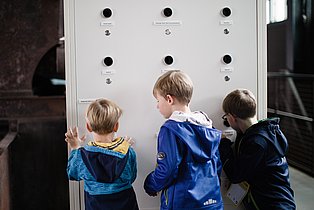 Drei Kinder stehen vor einer Entdeckerwand und schauen durch verschiedene Linsen.
