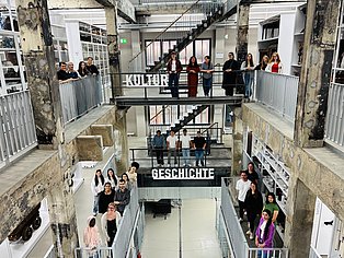 Menschen stehen auf drei Etagen eines offenen Industriegebäudes, das zu einem modernen Archiv oder Museumsdepot umgebaut wurde. Auf jeder Etage posieren Gruppen junger Erwachsener und blicken freundlich in die Kamera. Auf mittlerer Höhe sind die großen Wörter „KULTUR“ und darunter „GESCHICHTE“ an den Geländern angebracht. Die Umgebung ist geprägt von rauem Beton, industriellen Treppenkonstruktionen und offenen Regalsystemen mit Museumsobjekten, was auf ein kulturhistorisches Umfeld hinweist. Die Szene wirkt dynamisch, offen und gemeinschaftlich.