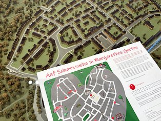 Gedruckte Schatzkarte mit Lageplan der Margarethenhöhe schwebt über einem Architekturmodell der Margarethenhöhe
