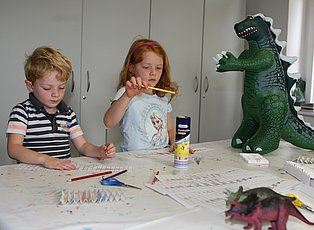 Zwei Kinder beim Malen und vor ihnen auf dem Tisch stehen kleine Dinos.