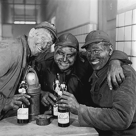 Three mountain farmers hang in each other's arms and hold bottles of "Schlegel Gold" beer in their hands.